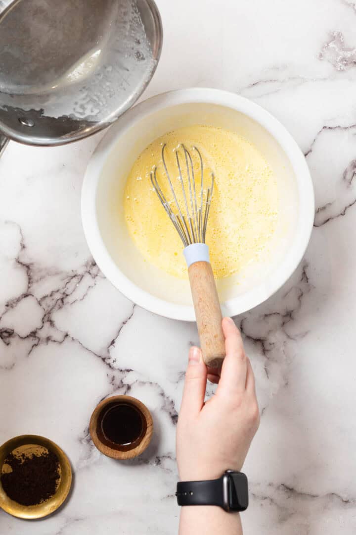 A hand holding a whisk rests in a bowl of beaten eggs on a marble countertop. Nearby are a pot with milk, a small bowl of vanilla extract, and a dish with ground coffee.