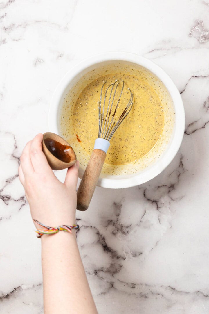 A hand holds a small wooden bowl, pouring liquid into a white bowl of yellow mixture with visible spices. A whisk rests in the bowl, and the person wears multicolored string bracelets. The background is a marble surface.