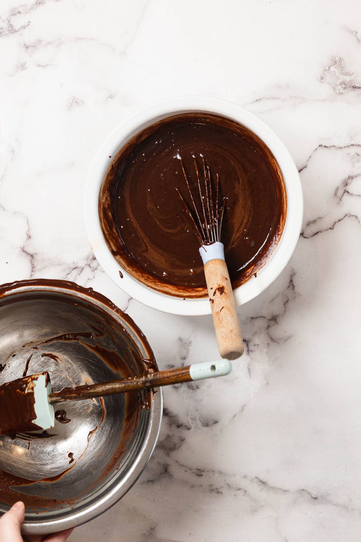 Two bowls on a marble surface; one white bowl with whisk, both containing chocolate batter, and one metal bowl with rubber spatula resting inside. A hand holds the metal bowl.
