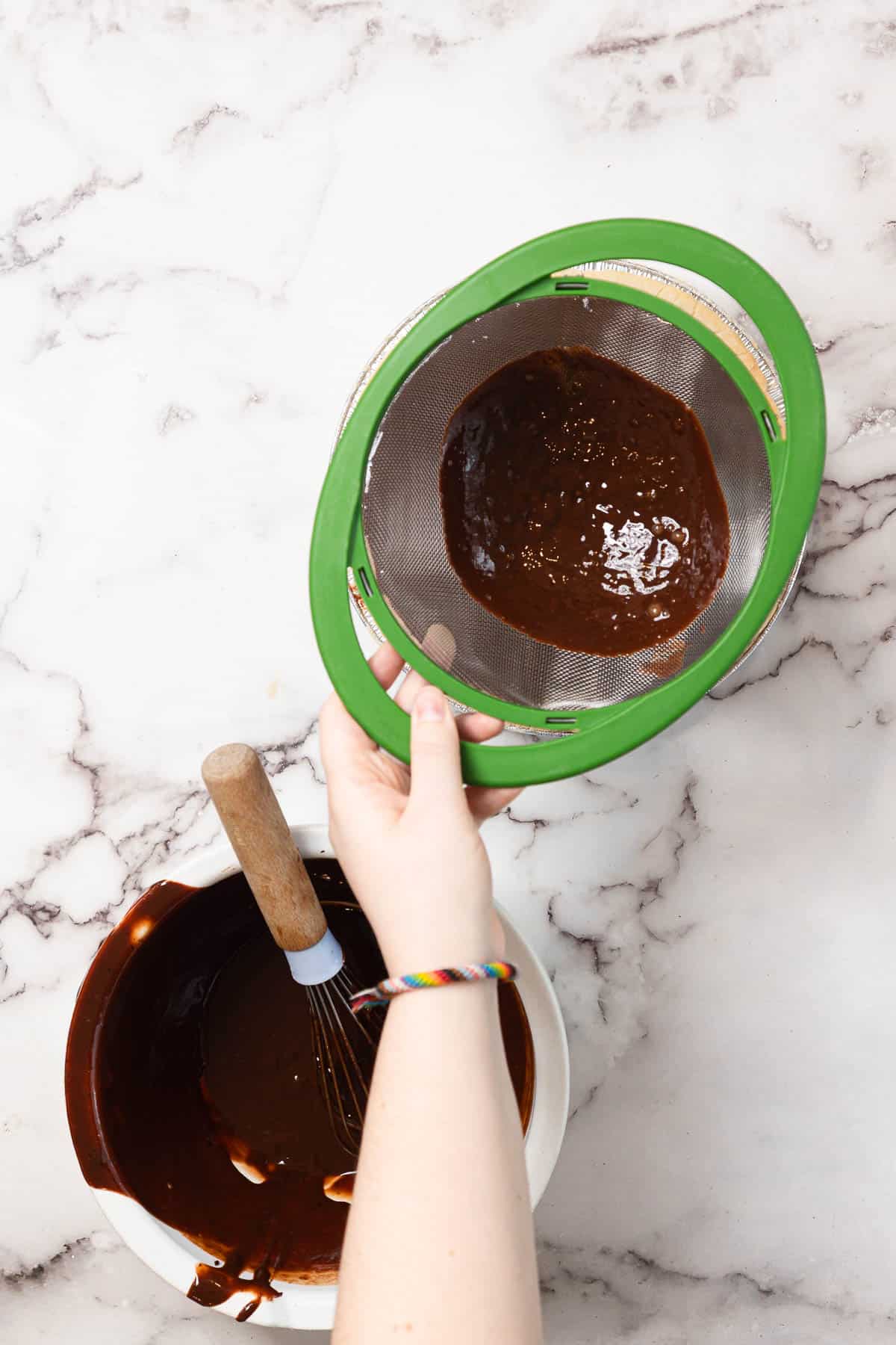 A person holds a green sieve with chocolate batter above a white bowl containing more chocolate batter and a whisk, on a marble countertop.