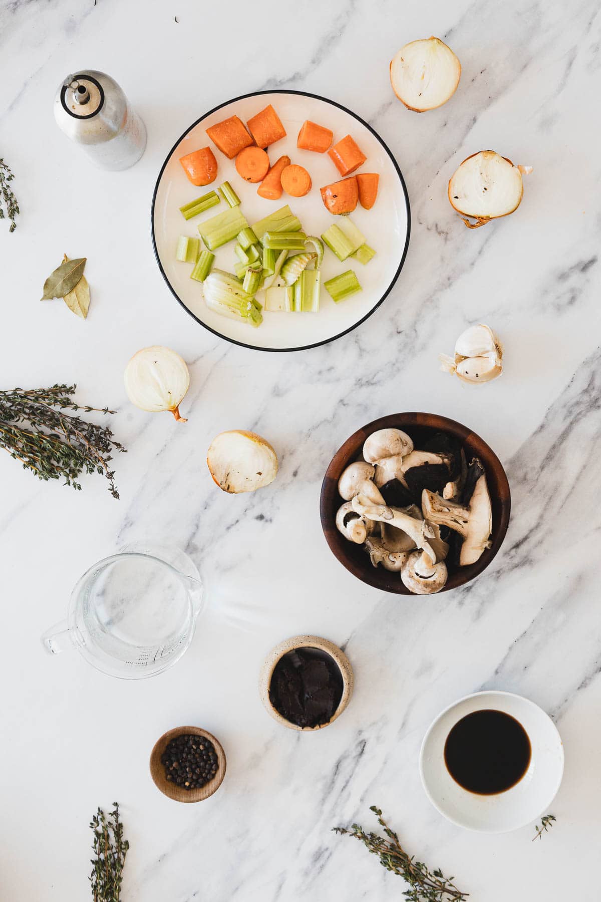 A flat lay of Roasted Mushroom Broth soup ingredients on a marble surface, including chopped carrots, celery, onions, mushrooms, a bowl of soy sauce, peppercorns, a bay leaf, fresh herbs, and a measuring cup of water.
