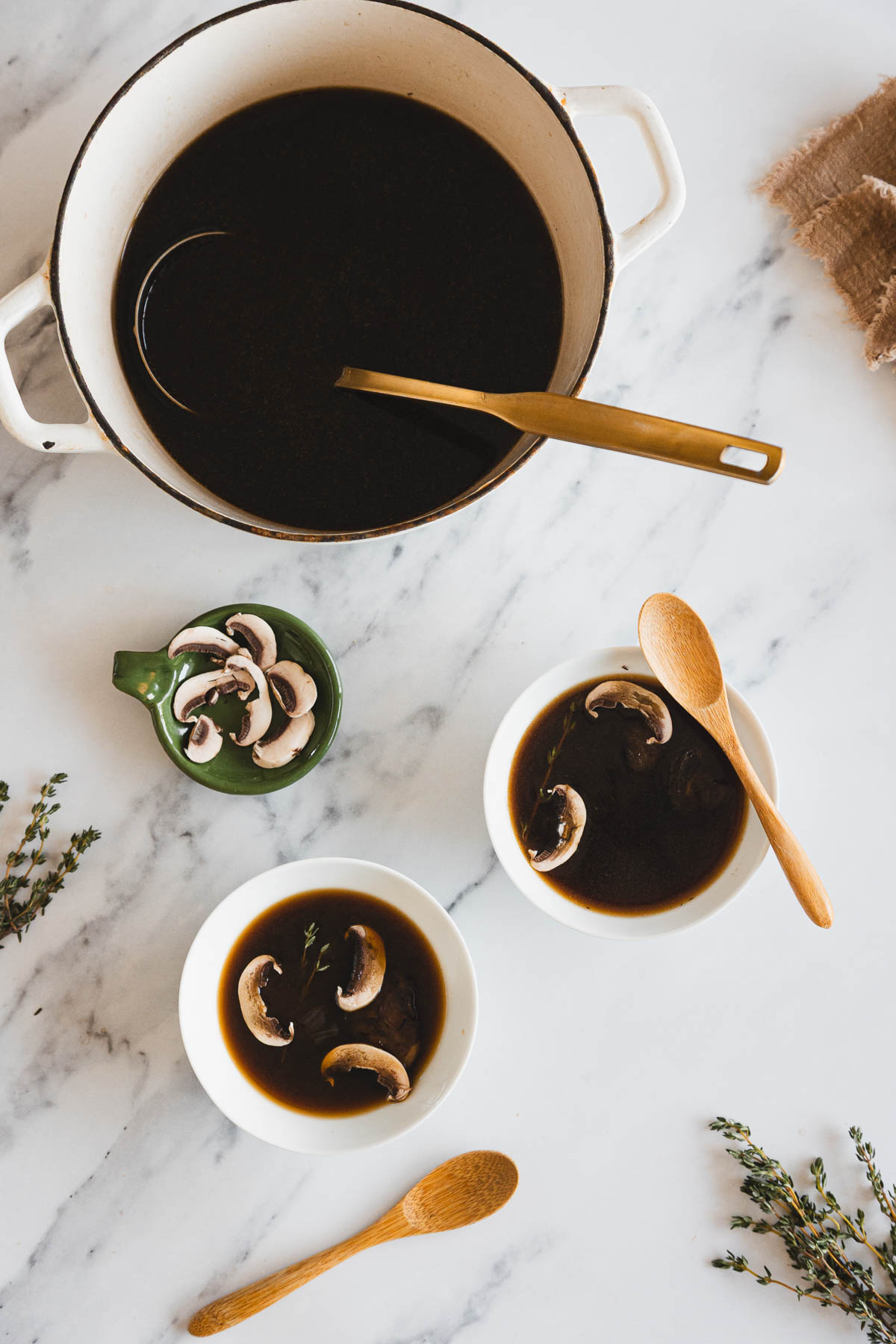 A pot of dark Roasted Mushroom Broth with a ladle sits on a marble surface. Nearby are two bowls of soup with mushroom slices and herbs, wooden spoons, loose thyme sprigs, and a small dish of raw mushroom slices.