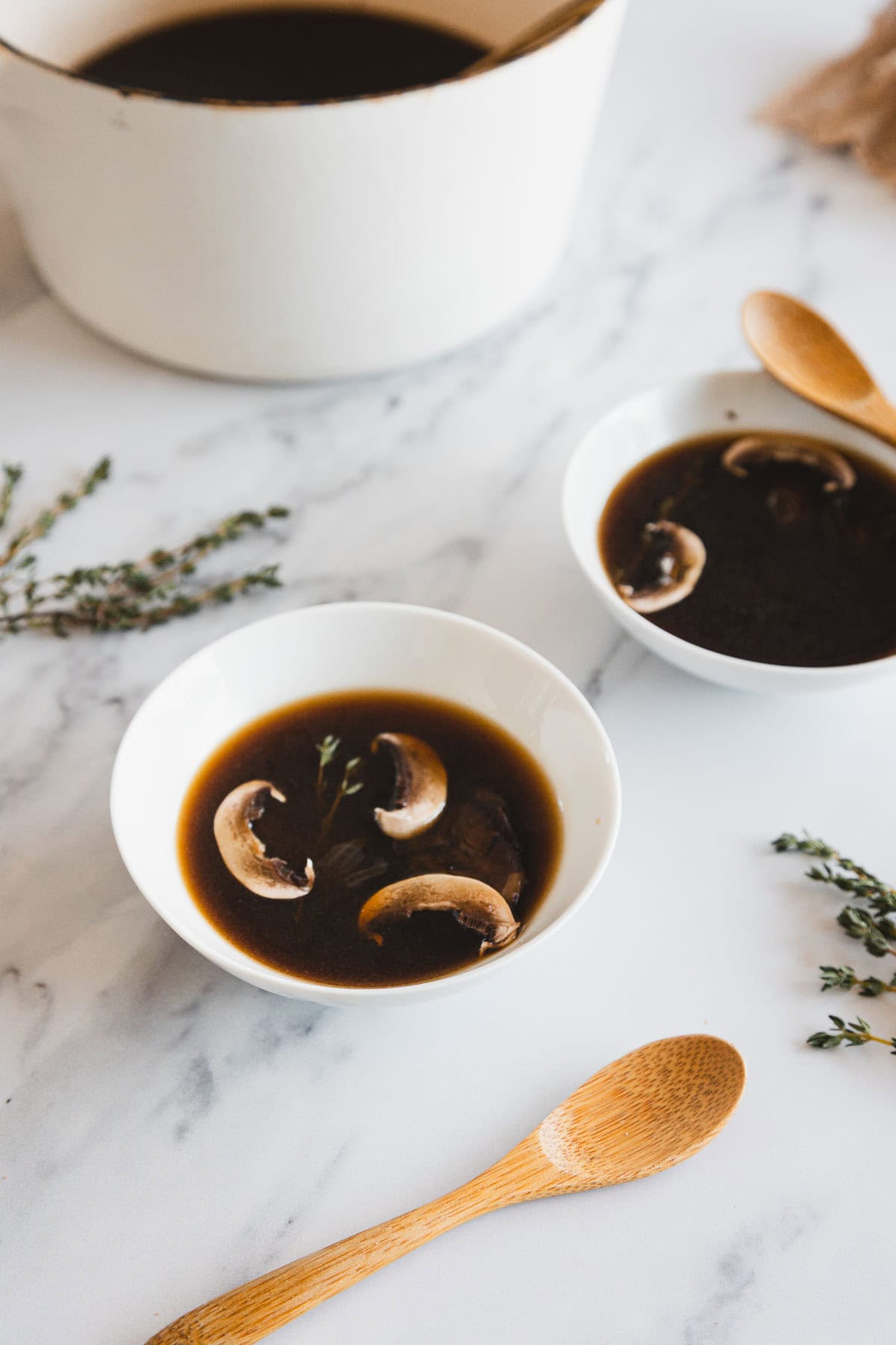 Two white bowls filled with roasted mushroom broth and mushroom slices sit on a marble surface, with a white pot in the background, sprigs of herbs, and wooden spoons nearby.