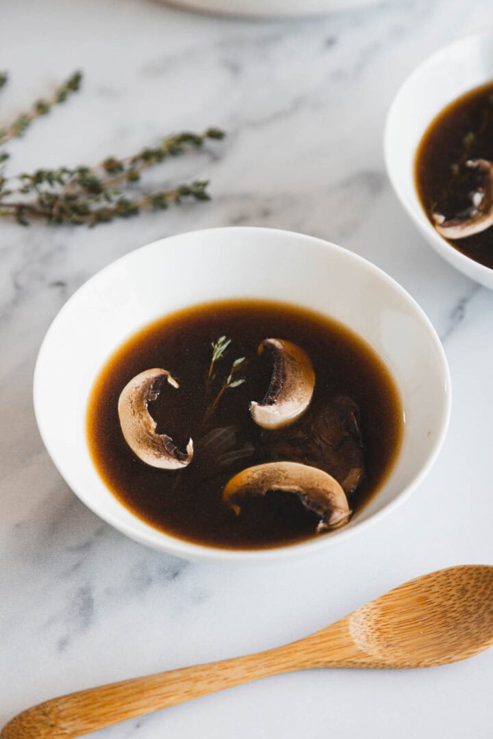 A white bowl filled with roasted mushroom broth, containing three mushroom slices and a small sprig of thyme, sits on a marble surface next to a wooden spoon and some fresh thyme.
