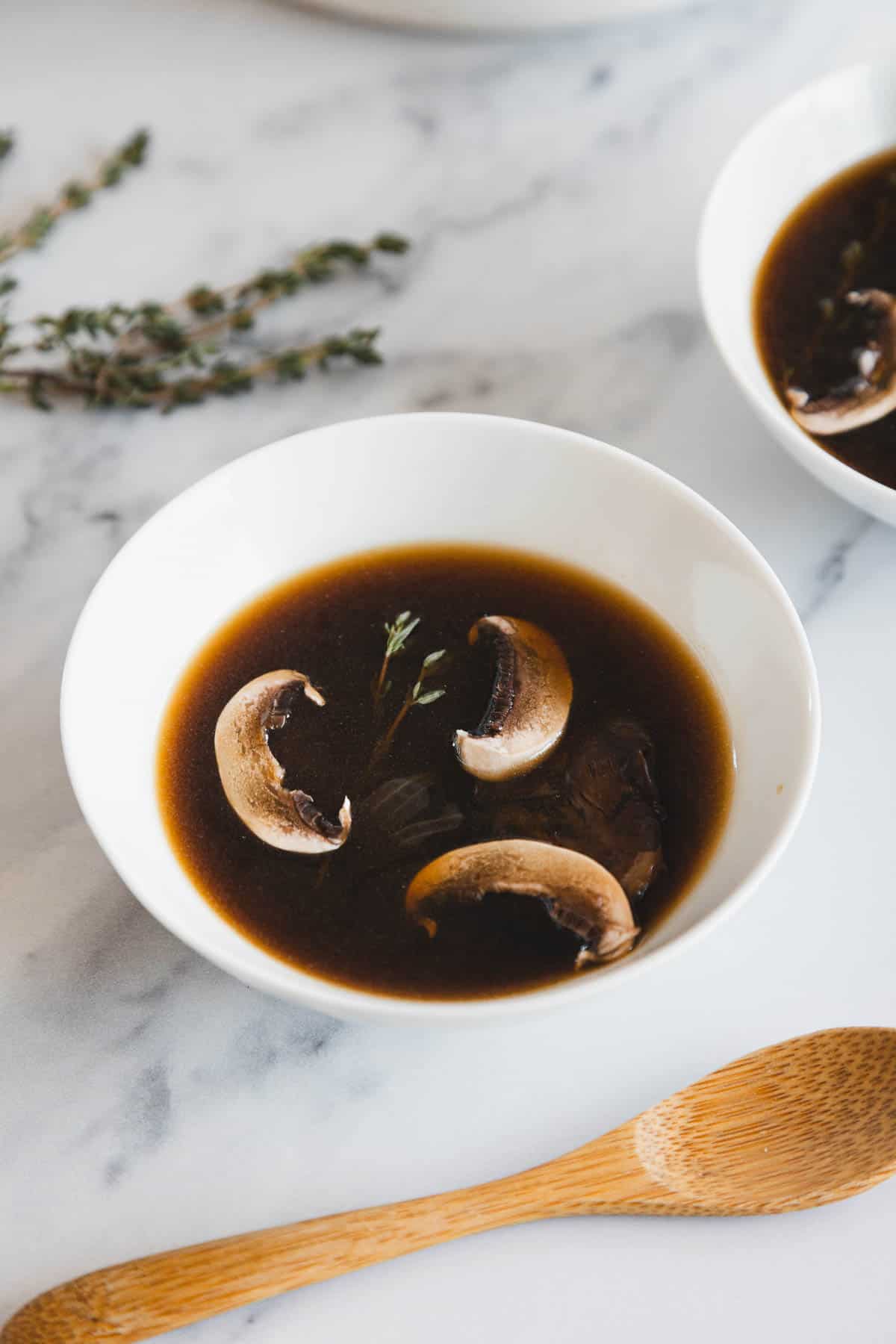 A white bowl filled with roasted mushroom broth, containing three mushroom slices and a small sprig of thyme, sits on a marble surface next to a wooden spoon and some fresh thyme.