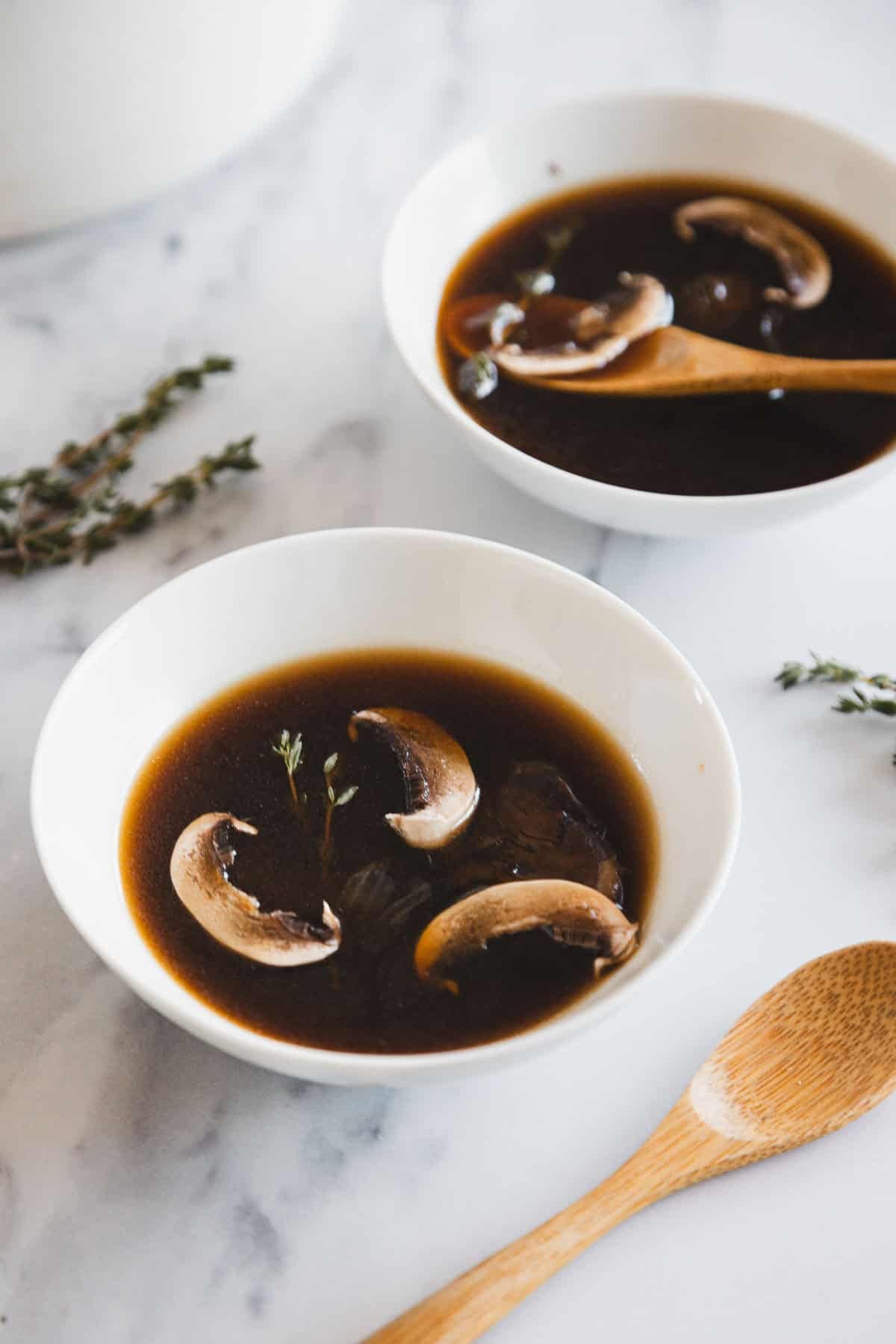 Two white bowls filled with Roasted Mushroom Broth and sliced mushrooms sit on a marble surface. A wooden spoon rests in one bowl, with sprigs of fresh thyme nearby. Another wooden spoon lies beside the bowls.