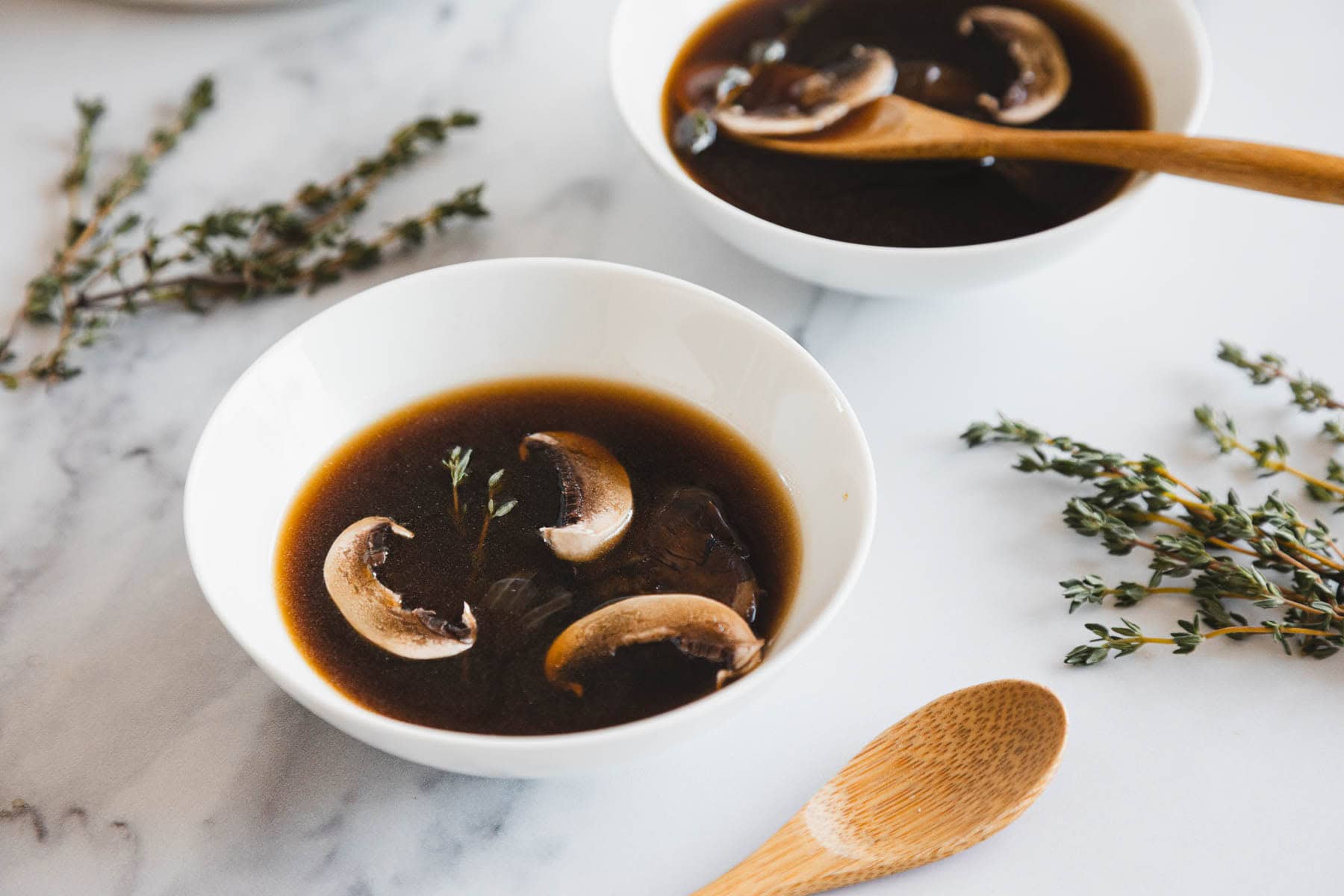 Two white bowls of roasted mushroom broth with mushroom slices and herbs sit on a marble surface, accompanied by wooden spoons and sprigs of fresh thyme nearby.