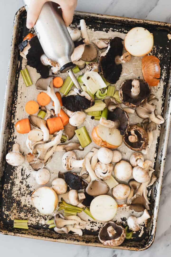 A hand holds an oil dispenser over a baking sheet filled with assorted raw vegetables, including carrots, celery, onions, and various mushrooms&mdash;perfect ingredients for a rich Roasted Mushroom Broth&mdash;on a marble surface.