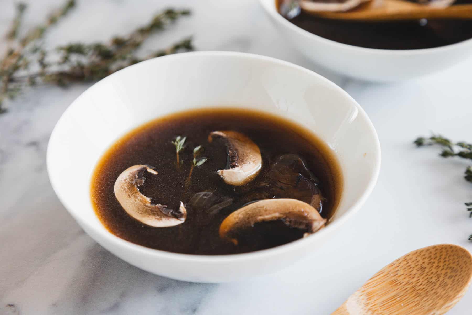 A white bowl filled with roasted mushroom broth and several sliced mushrooms, garnished with a sprig of thyme, sits on a white surface next to a wooden spoon and some scattered herbs.