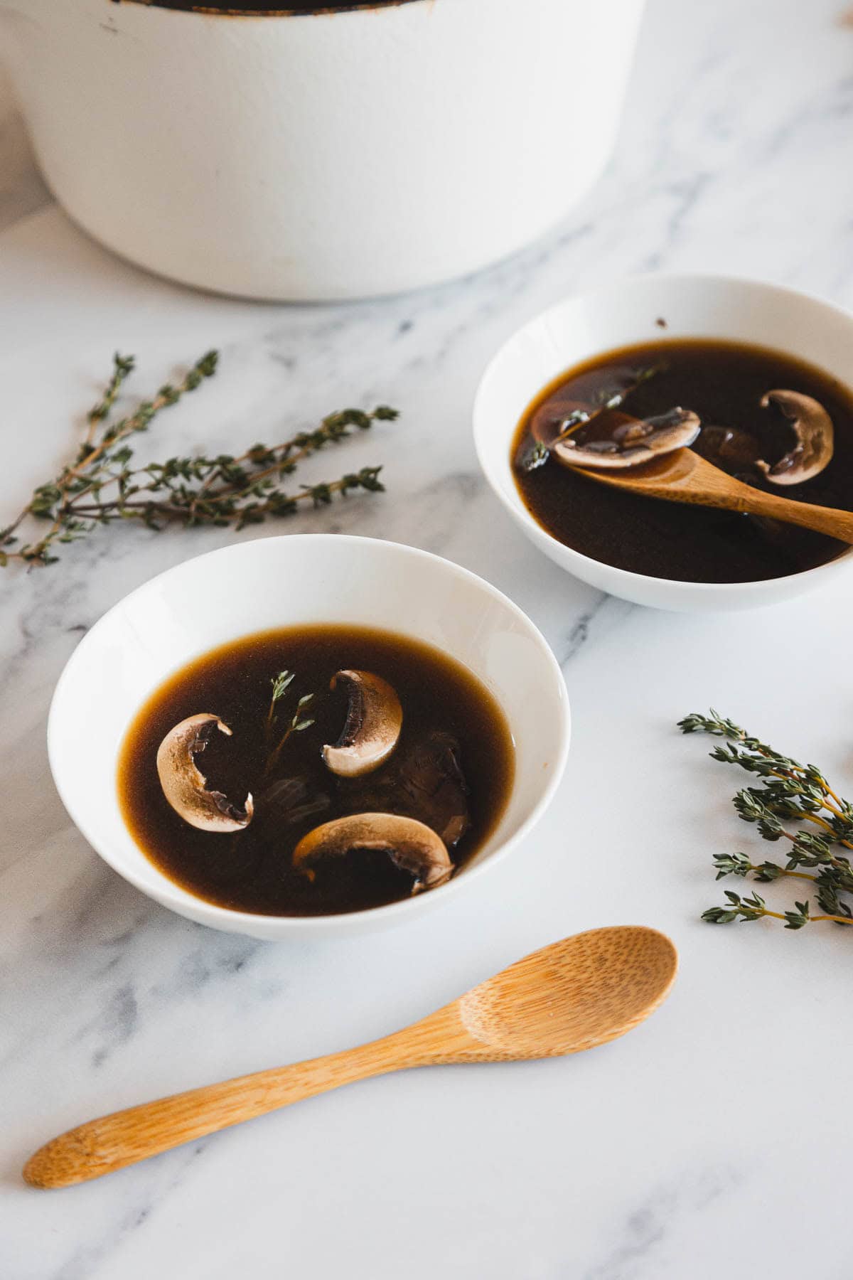 Two white bowls of Roasted Mushroom Broth with sliced mushrooms and thyme sprigs sit on a white marble surface. A wooden spoon rests near the bowls, with fresh thyme sprigs artfully arranged around them.