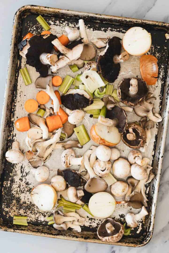A metal baking sheet with raw, chopped vegetables including mushrooms, carrots, celery, and onions spread in an uneven layer&mdash;perfect for making a rich Roasted Mushroom Broth. The baking sheet shows visible stains and scratches.