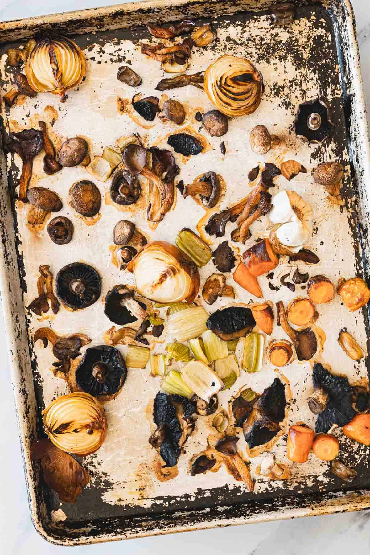 A metal baking sheet with roasted vegetables, including whole onions, sliced carrots, celery pieces, garlic cloves, and various mushrooms&mdash;perfect ingredients for Roasted Mushroom Broth&mdash;spread irregularly across its stained and slightly worn surface.