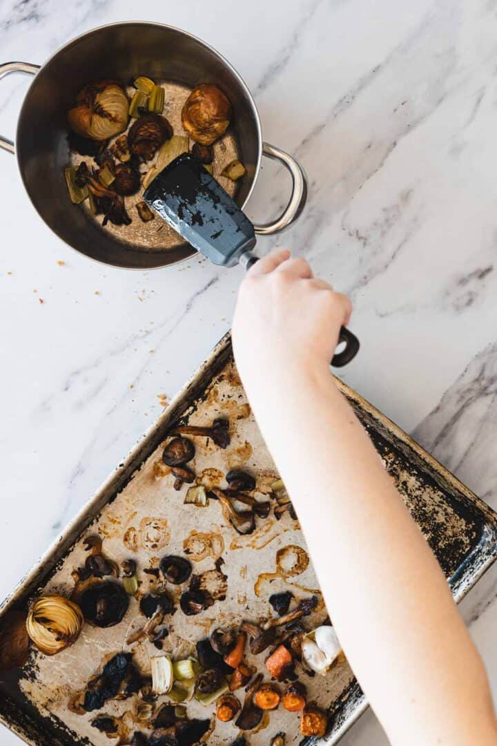 A person uses a black spatula to transfer roasted vegetables for Roasted Mushroom Broth from a baking sheet lined with parchment paper into a metal pot on a marble countertop.