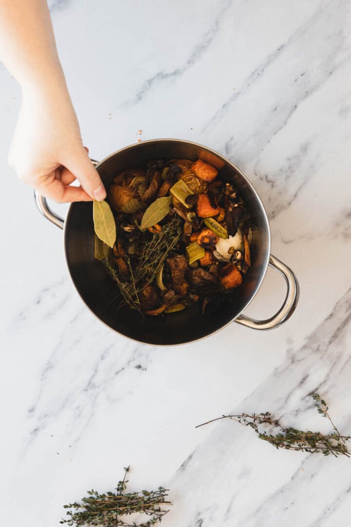 A hand is placing a bay leaf into a pot filled with chopped vegetables and herbs, including carrots, celery, thyme, and mushrooms&mdash;perfect ingredients for a flavorful Roasted Mushroom Broth&mdash;on a white marble surface.