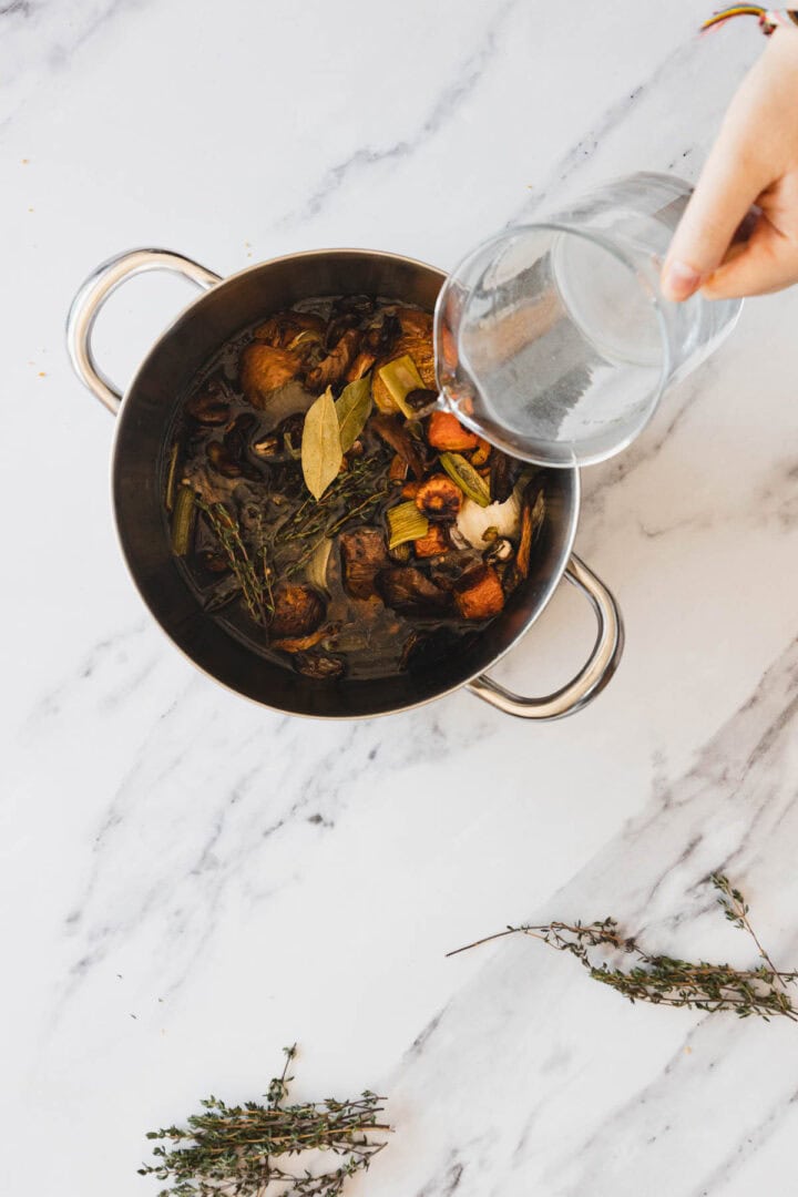 A hand pours water from a glass pitcher into a stainless steel pot containing herbs, bay leaves, mushrooms, and other vegetables to prepare Roasted Mushroom Broth, all placed on a white marble surface.