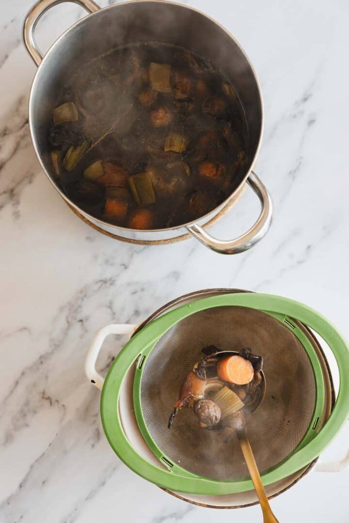 A pot of simmered vegetables in dark Roasted Mushroom Broth sits beside a mesh strainer over a bowl. A slotted spoon rests in the strainer, holding pieces of cooked carrot and other vegetables.