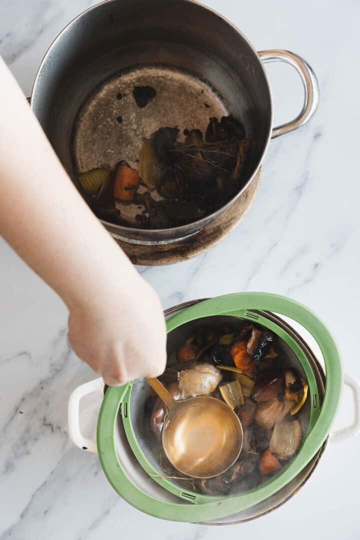 A hand uses a ladle to strain Roasted Mushroom Broth through a mesh sieve into a white pot, separating liquid from cooked vegetable scraps, with an empty metal pot nearby on a marble surface.