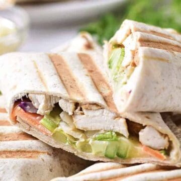 Two grilled wraps cut in half, showing visible fillings of chicken, cucumbers, shredded lettuce, carrots, and sauce, placed on a plate with blurred greens and a bowl in the background.