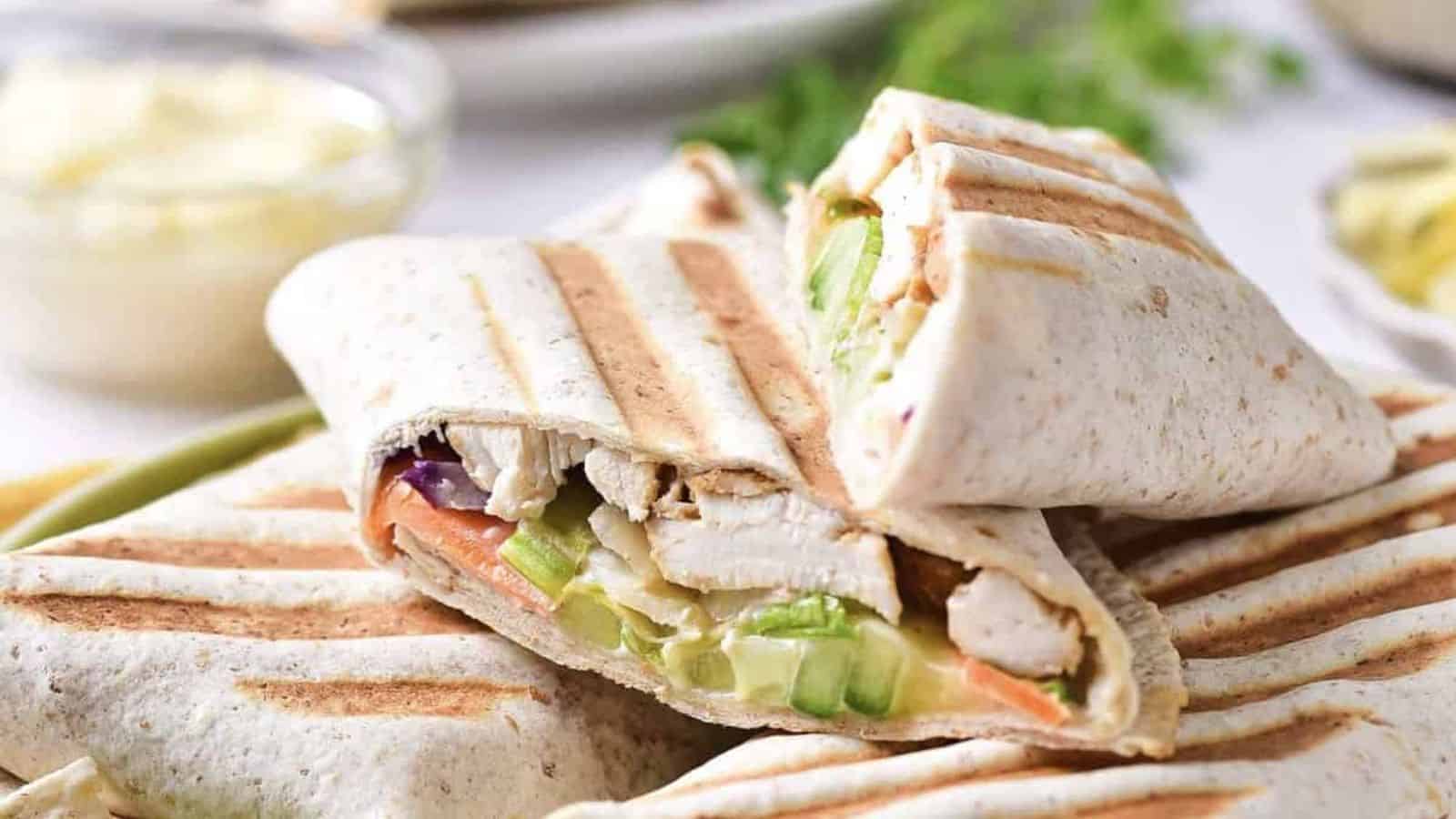 Two grilled wraps are cut in half, showing fillings of grilled chicken, lettuce, cucumber, and other vegetables. The wraps have visible grill marks and are stacked on a plate with a blurred background.