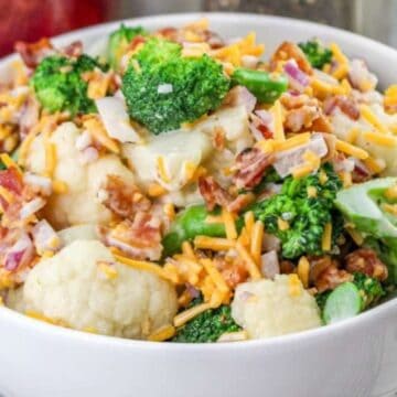 A white bowl filled with a salad made of cauliflower, broccoli, shredded cheddar cheese, bacon bits, and chopped red onions, set on a checkered cloth next to a spoon.