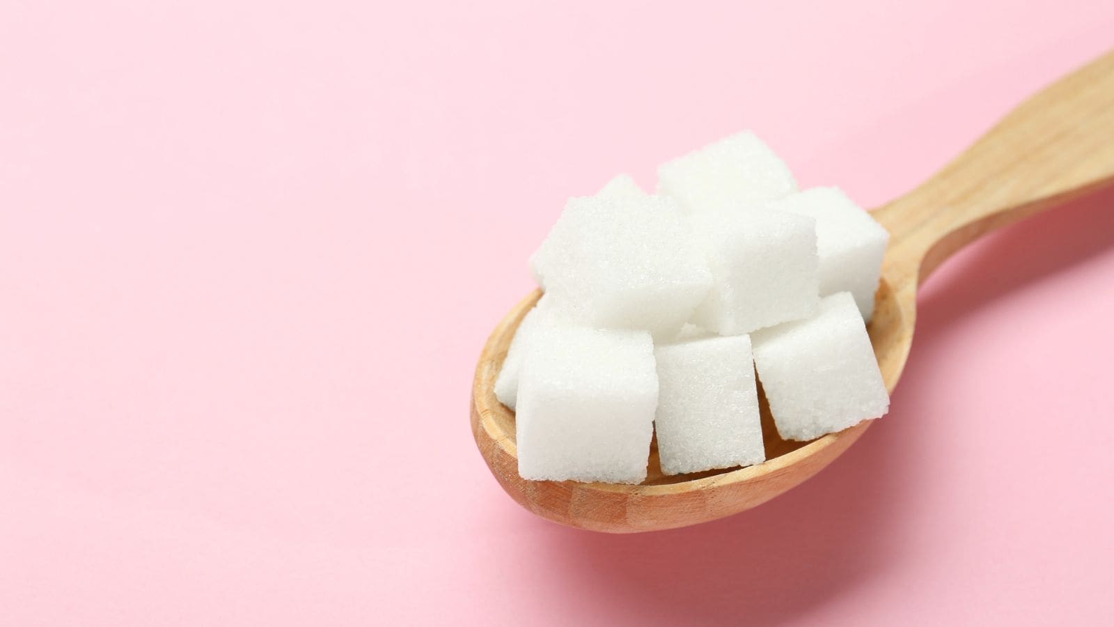 A wooden spoon holding several white sugar cubes is placed against a plain light pink background.