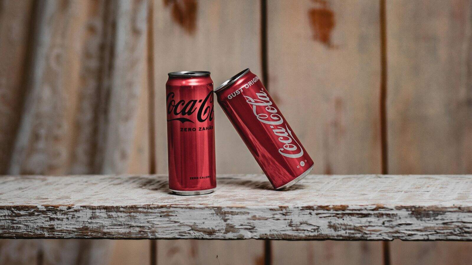 Two red Coca-Cola cans are placed on a rustic wooden surface in front of a light brown wooden background. One can is upright and labeled "Zero Zahar," while the other leans at an angle.