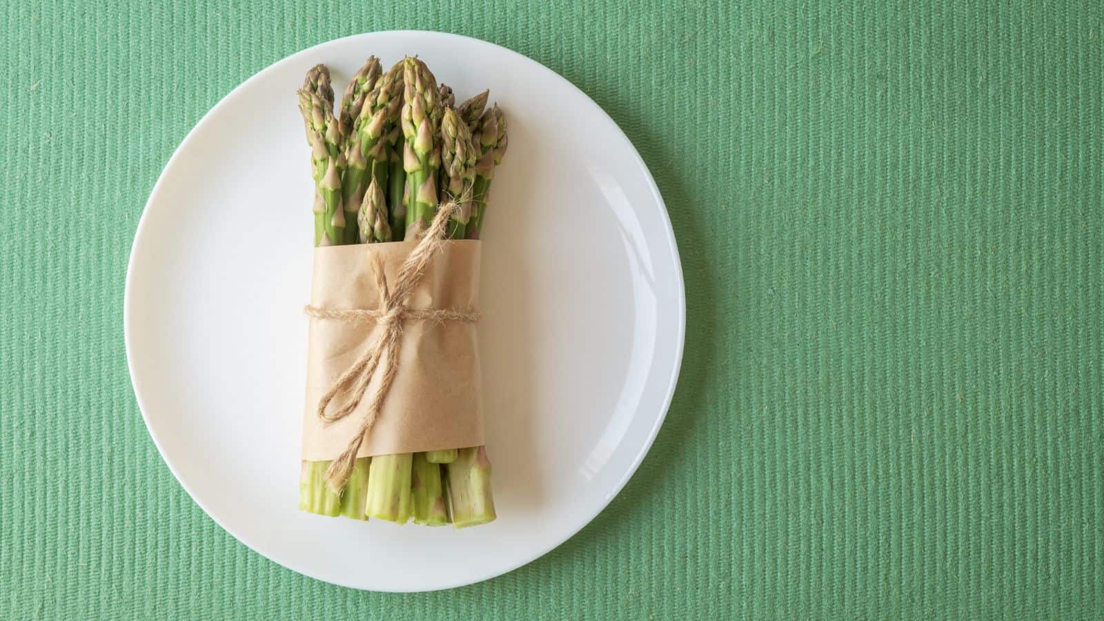 A bundle of asparagus spears wrapped in brown paper and tied with string is placed on a white plate, which sits on a green textured surface.