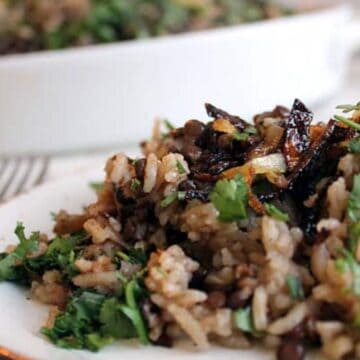 A plate of rice and lentils topped with chopped herbs and caramelized onions is served in the foreground, with a casserole dish of the same dish in the background.