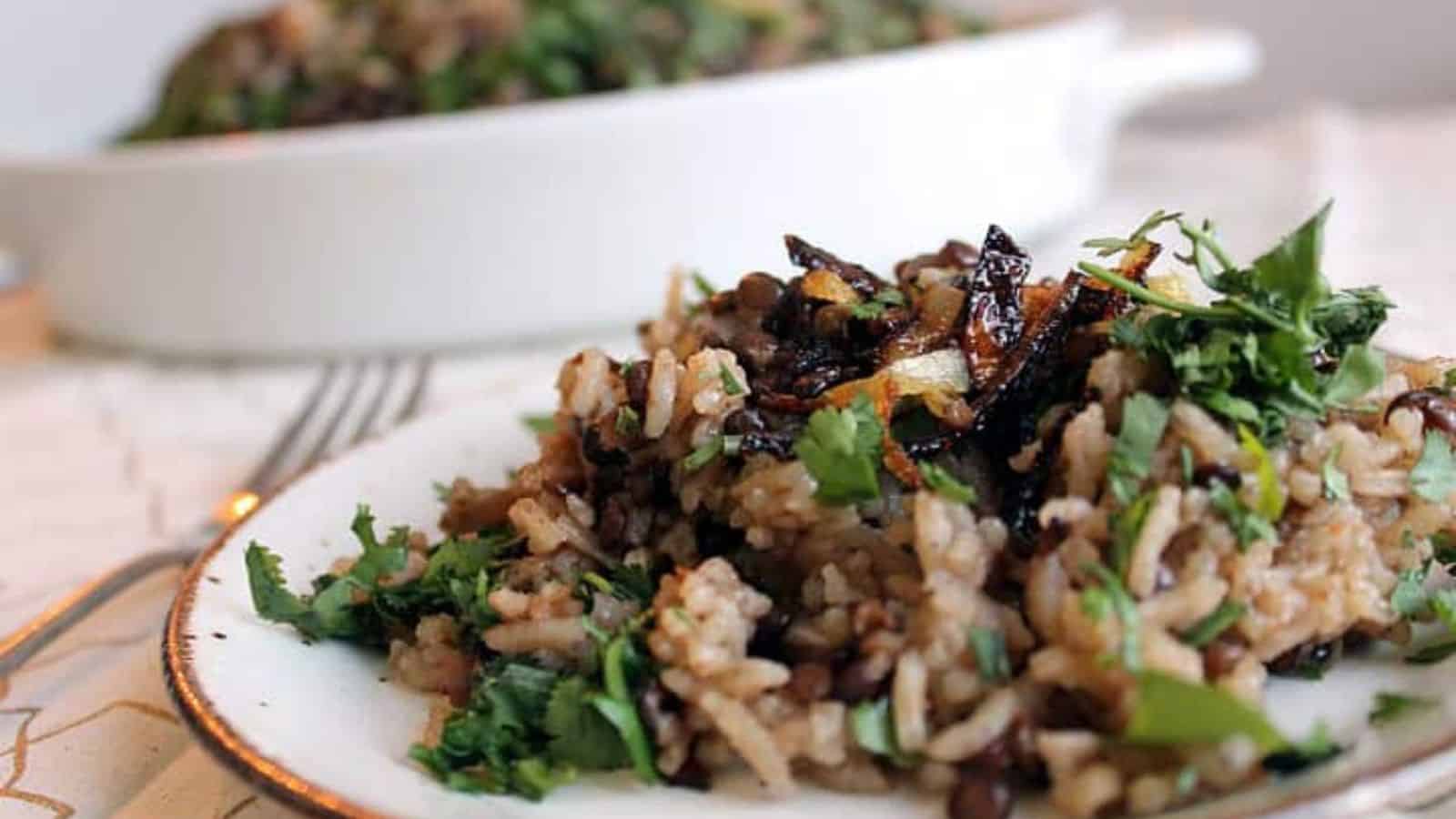 A white dish filled with cooked lentils and rice, topped with chopped cilantro and caramelized onions. A wooden spoon rests on a patterned napkin next to the dish.