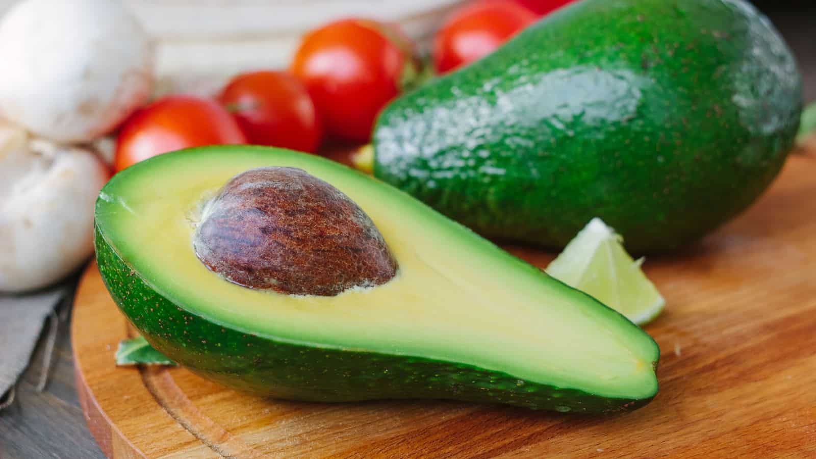 A halved avocado with pit, next to a whole avocado, lime wedge, tomatoes, and mushrooms on a wooden cutting board.
