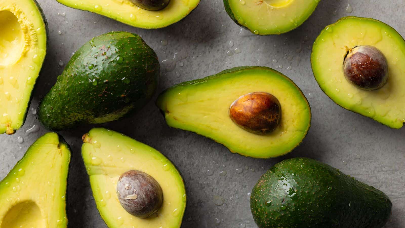 A group of fresh avocados, some whole and some halved, are arranged on a gray surface. The avocado halves reveal their seeds and yellow-green flesh with visible water droplets.