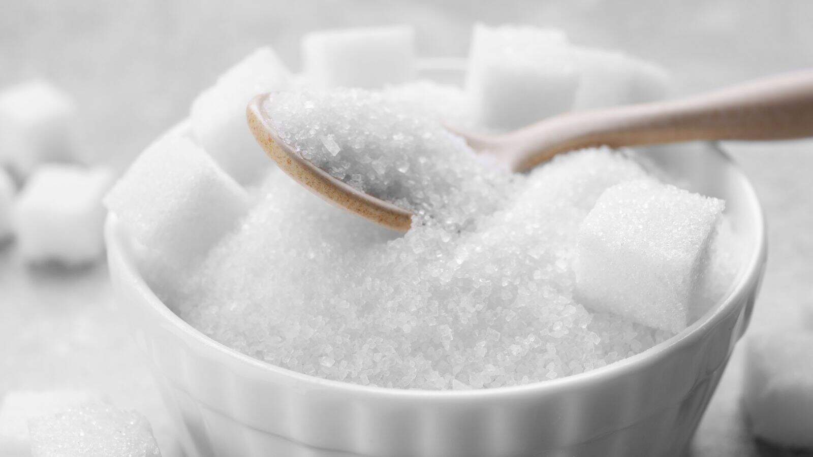 A white ceramic bowl filled with granulated sugar and several sugar cubes. A wooden spoon rests on top of the sugar, with some granules on the spoon. The background is softly blurred.