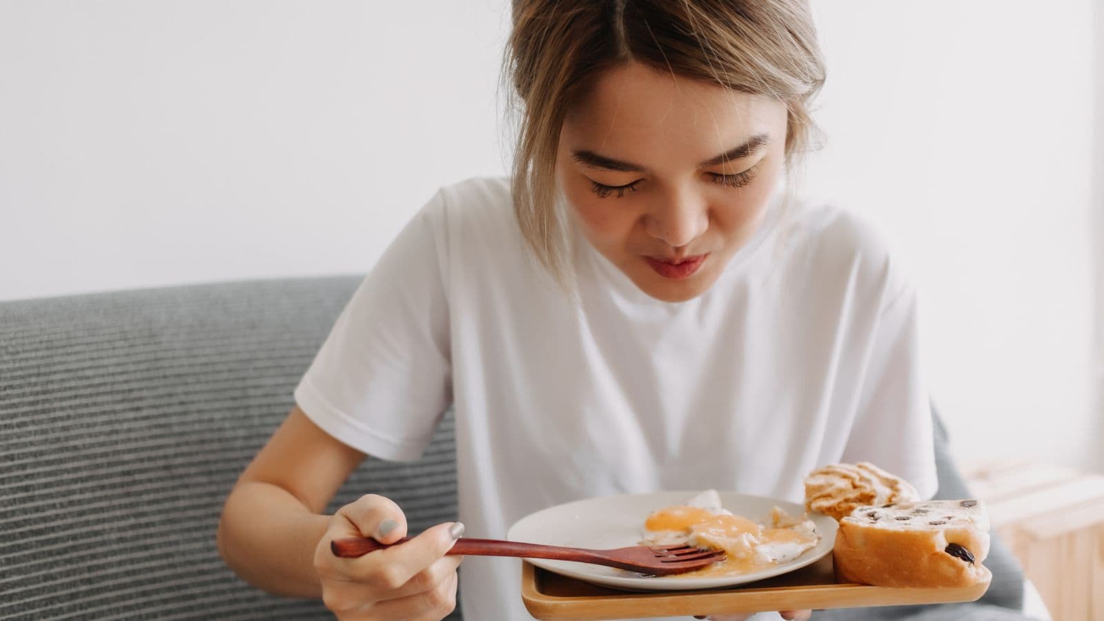 A person wearing a white shirt is sitting on a gray chair, looking down at a plate of food with a pastry and a cooked egg, using a fork and knife.