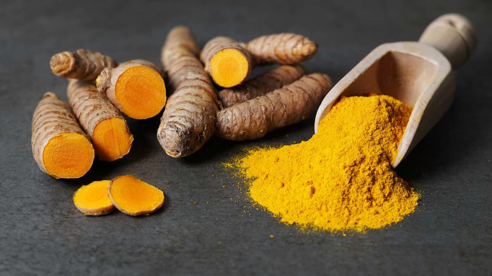 Whole turmeric roots, some cut open to show their bright orange interior, are arranged on a dark surface next to a wooden scoop filled with vibrant yellow turmeric powder.