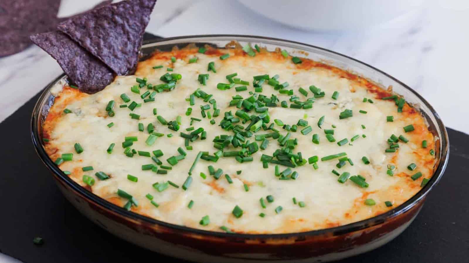 A round glass dish filled with a baked dip topped with melted cheese and chopped chives, with two blue corn tortilla chips placed upright in the dip.