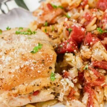A seasoned baked chicken thigh topped with grated cheese is served next to orzo pasta mixed with diced tomatoes and herbs on a white plate. A fork and knife are partially visible beside the food.