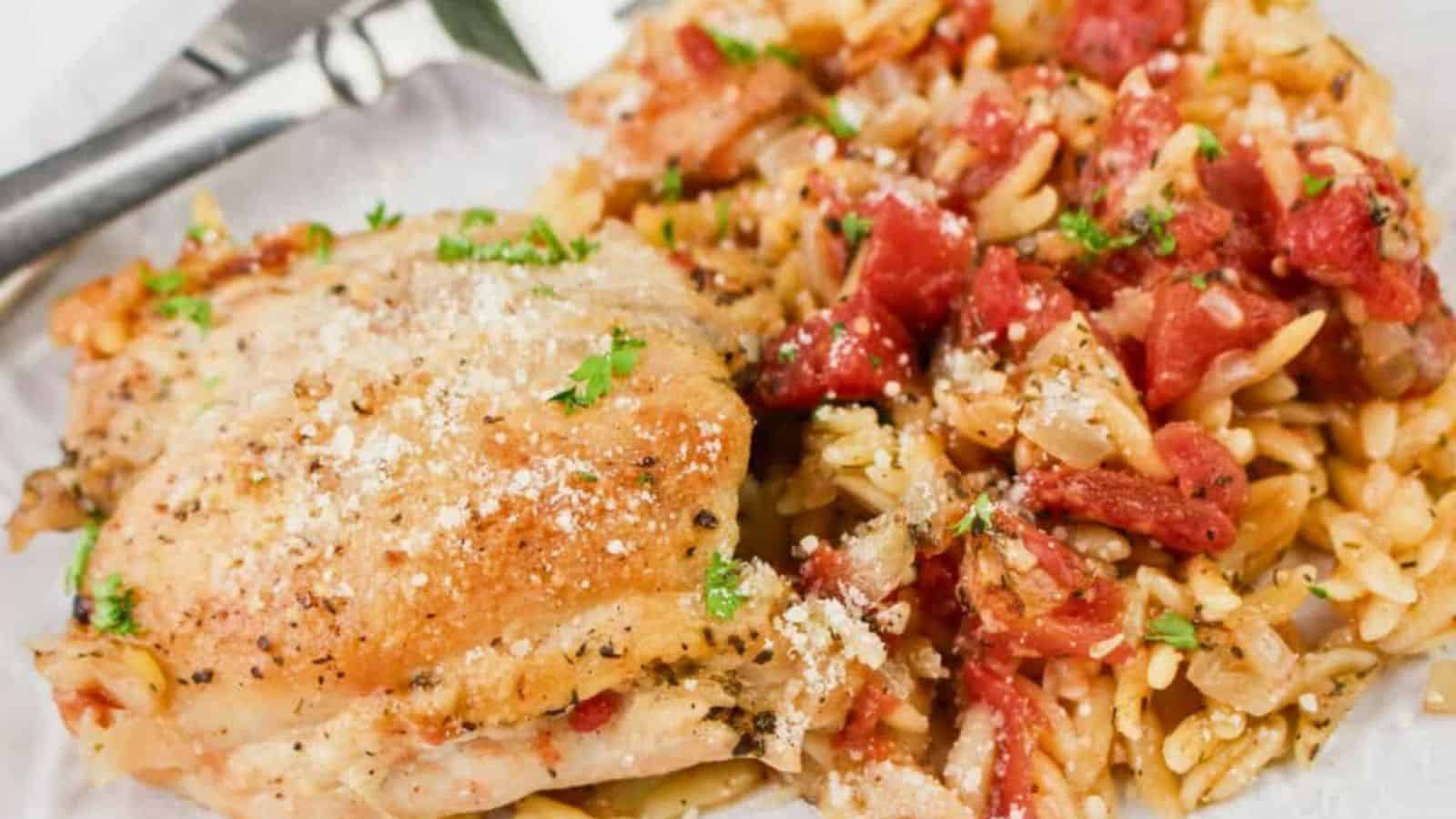 A plate with baked chicken thigh and orzo pasta mixed with tomatoes, garnished with herbs, with a fork and knife beside.