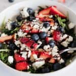 A bowl of Balsamic Berry Salad with Goat Cheese features mixed greens, sliced strawberries, blueberries, crumbled goat cheese, and a drizzle of dark balsamic glaze, with a fork resting inside the bowl.