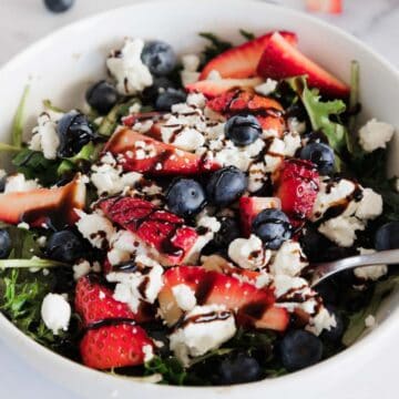 A bowl of Balsamic Berry Salad with Goat Cheese features mixed greens, sliced strawberries, blueberries, crumbled goat cheese, and a drizzle of dark balsamic glaze, with a fork resting inside the bowl.