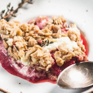 A serving of fruit crumble with a crumbly oat topping and a layer of red fruit filling, garnished with a sprig of thyme on a white plate with a brown rim, next to a silver spoon.
