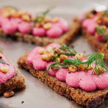 Four rectangular pieces of brown bread topped with pink spread, toasted pine nuts, and fresh dill are arranged on a metal tray.