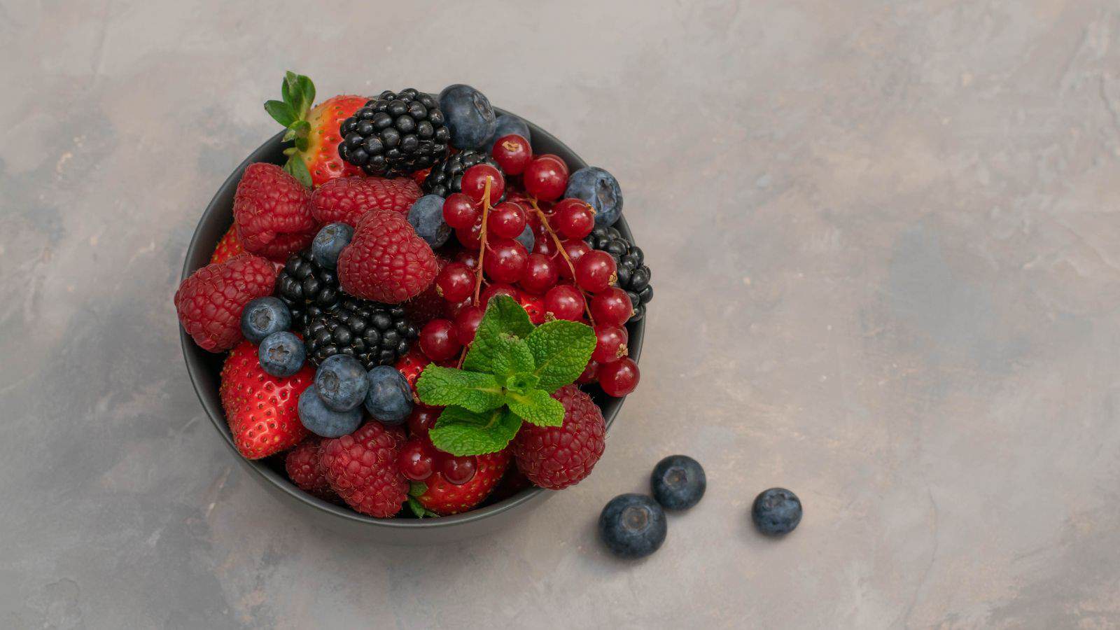 A black bowl filled with assorted fresh berries including strawberries, raspberries, blueberries, blackberries, and red currants, garnished with mint leaves, sits on a gray textured surface with a few blueberries beside it.