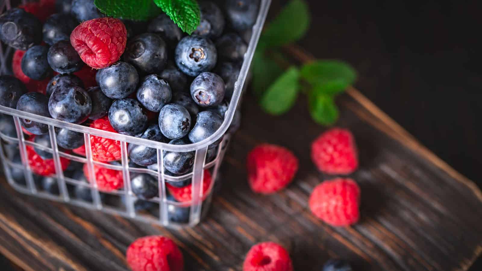 A clear plastic container filled with fresh blueberries and a few raspberries, sits on a wooden surface with several loose raspberries and green mint leaves scattered around.