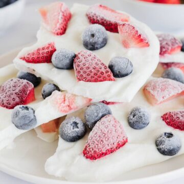 A plate of frozen yogurt bark topped with sliced strawberries and whole blueberries. The bark pieces are white with visible frost, and bowls of additional berries are blurred in the background.