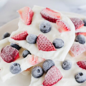 Several pieces of white frozen yogurt bark topped with whole blueberries and sliced strawberries are stacked on a white plate.