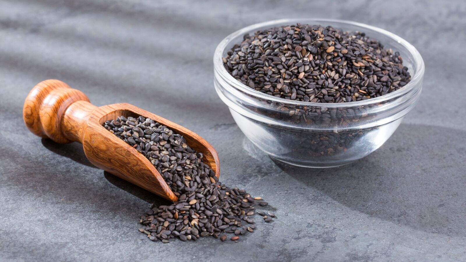 A glass bowl filled with black sesame seeds sits on a gray surface next to a wooden scoop, which also holds some black sesame seeds, with a few seeds scattered nearby.