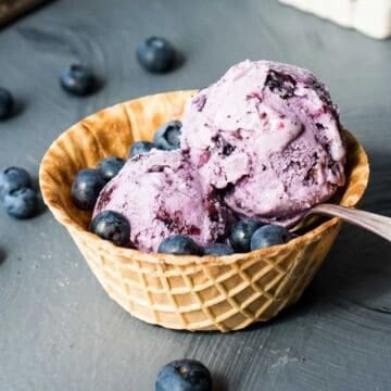 A waffle bowl filled with two scoops of blueberry ice cream, garnished with fresh blueberries, sits on a gray surface with additional blueberries scattered around. A spoon rests in the bowl.
