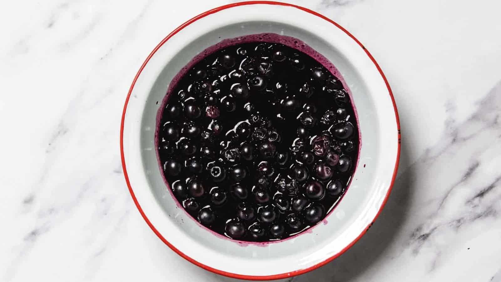 A white bowl with a red rim contains a serving of dark purple berries in liquid, placed on a white marble surface.