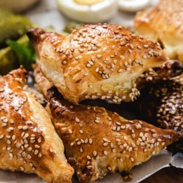 A close-up of golden-brown, triangular puff pastries topped with sesame seeds, stacked on parchment paper, with pickles and olives visible in the background.