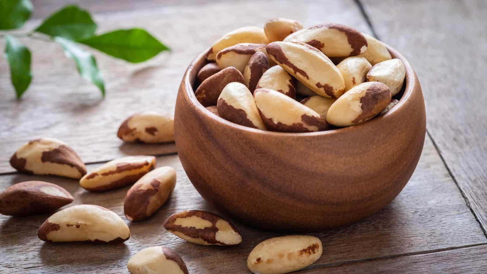 A wooden bowl filled with Brazil nuts sits on a wooden surface, with a few Brazil nuts scattered around the bowl. Green leaves are visible in the background.